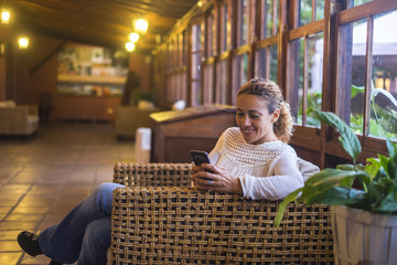 woman sitting on wicker chair in wooden house with large picture window smiling looking on cellphone. Tehcnlogy leisure activity for cheerful people indoor