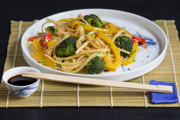 Asian noodles with vegetables and soy sauce on bamboo mat and chopsticks, black background