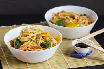 Asian food, two bowls of noodles with vegetables and soy sauce on a bamboo mat, black background