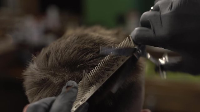 Male hands dressed in black gloves is cutting short hair of the client with the help of comb in a barbershop