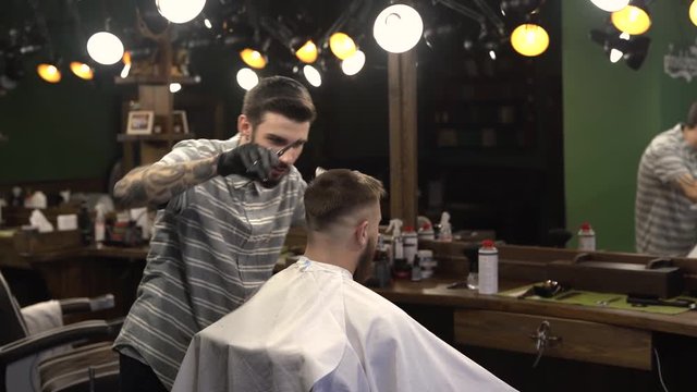 Young man with tattoos and brown beard dressed in a striped T-shirt is drying the  hair of satisfied client in the barbershop.There is backlighting around the room in a retro style.