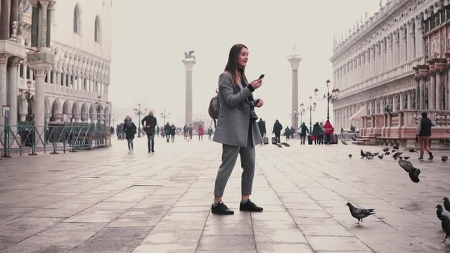 Happy smiling tourist girl takes smartphone photos of pigeons flock standing on city square in Venice, Italy slow motion