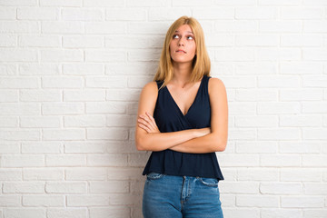 Blonde woman having doubts and with confuse face expression while bites lip. Questioning an idea on white brick wall background
