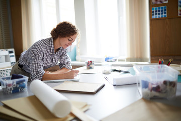 Busy young woman making notes or sketching in notepad while leaning over workplace
