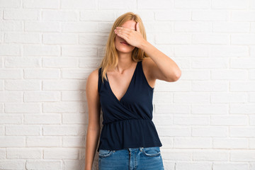 Blonde woman covering eyes by hands. Do not want to see something on white brick wall background