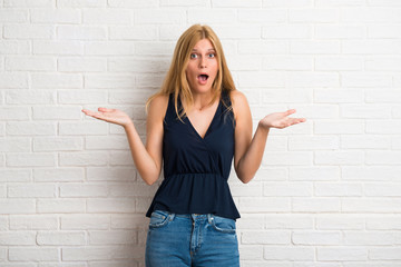 Blonde woman with surprise and shocked facial expression. Mouth open because not expect what has happened on white brick wall background