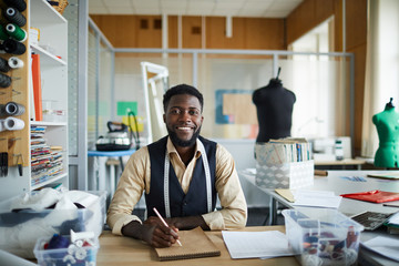 Young cheerful fashion designer sitting by table in his workshop and making new sketches in notepad