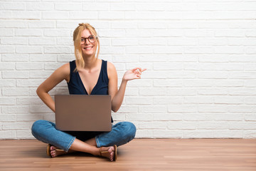 Blonde girl sitting on the floor with her laptop pointing finger to the side and presenting a product on white brick wall background