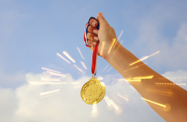 woman hand raised, holding gold medal against sky. award and victory concept.