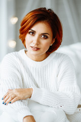 Portrait of a beautiful young girl with red hair in a white warm sweater. Close up. Woman posing and looking at the camera.