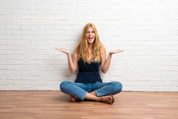 Blonde girl sitting on the floor with surprise and shocked facial expression on white brick wall background