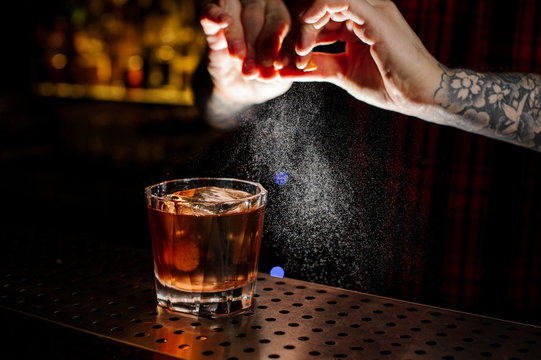 Tattooed bartender sprinkling orange peek juice into cocktail glass with whiskey