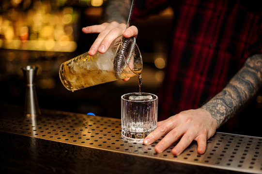 Barman pouring strong alcohol into a cocktail glass with ice cube using strainer