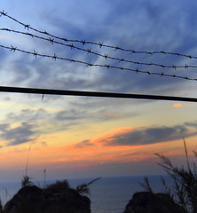 Trügerische Idylle: Stacheldrahtzaun vor Sonnenaufgangshimmel am Meer