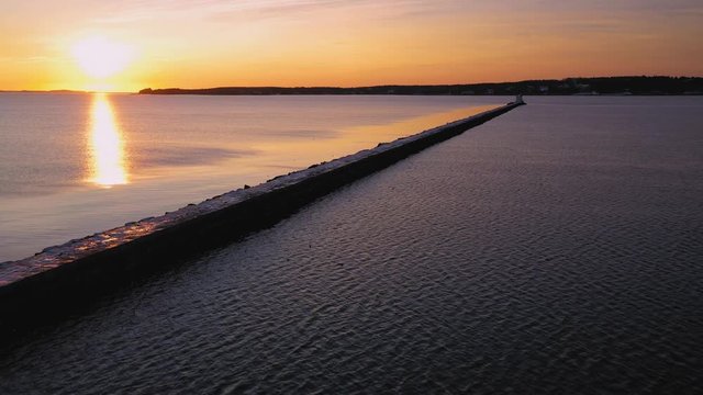 Aerial footage flying backwards at sunrise on the Maine coast away from a long pier with a lighthouse at the end of it
