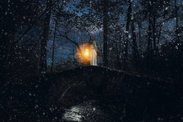 Girl in white dress with yellow light in her hand viewed from distance at night standing alone on hill in dark forest