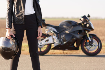 Cropped shot of unrecognizable woman biker dressed in black sportsclothes, holds protective helmet...