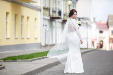 Naklejka premium young beautiful bride girl with wedding bouquet of roses stands on the street. the wind blows the veil