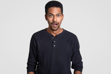 Indignant stupefied dark skinned young guy opens mouth with surprisement, dressed in casual jumper, reacts on sudden news from interlocutor, isolated over white background. Masculinity concept