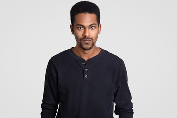 Obraz premium Indoor shot of serious self confident dark skinned young man with curly short hair, looks with stern expression, models indoor against white background. People, ethnicity and lifestyle concept