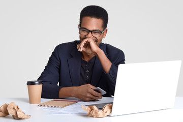 Photo of positive dark skinned man dressed in formal clothes, happy to read funny text message, holds modern cell phone, wears glaasses, surrounded with laptop computer, notepad, crags of paper