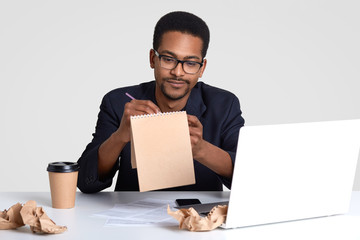 Shot of serious black man journalist works on creation new article, writes down ideas in spiral...