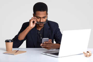 Surprised black man holds mobile phone, reads text message with astonishment, looks through spectacles, makes records in notepad, drinks takeaway coffee, poses at coworking space, isolated on white