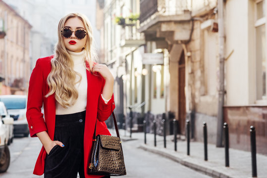 Outdoor fashion portrait of young beautiful fashionable lady wearing stylish glasses, white turtleneck, red blazer, holding stylish faux reptile skin bag, posing in street of european city. Copy space