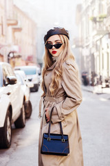 Obraz premium Outdoor fashion portrait of young beautiful fashionable girl wearing trendy beige long trench coat, stylish cat eye sunglasses, leather beret, holding handbag, model posing in street of european city