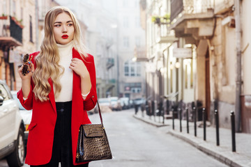 Outdoor fashion portrait of young beautiful fashionable woman wearing white turtleneck, red blazer,...