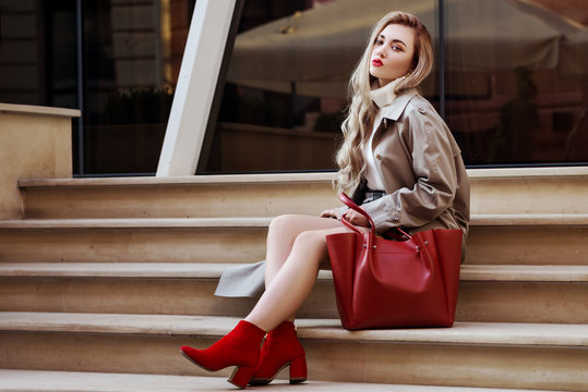 Outdoor fashion portrait of young beautiful fashionable woman wearing trendy beige long trench coat, red suede ankle boots, heels, holding big tote bag, sitting on stairs. Copy, empty space for text
