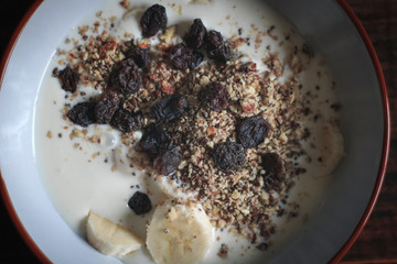 Vegan yogurt dish with pieces of banana, granola, raisins and oats on a dark wooden board illuminated with natural light of day