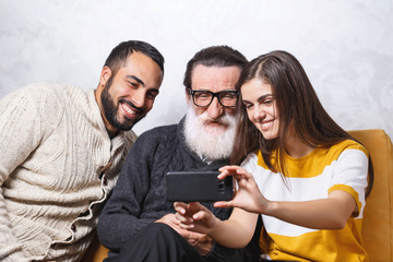 Brunette bearded son and brunette long haired daughter taking selfie with they senior bearded father in glasses while sitting on the yellow sofa in the light living room, modern technology