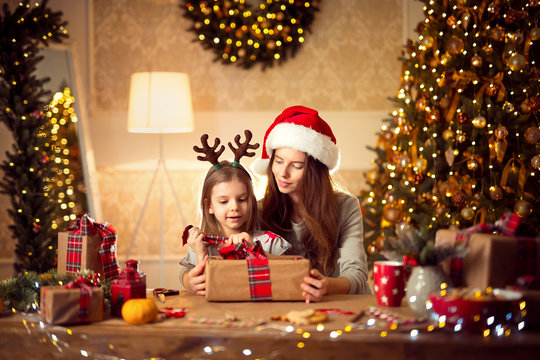 Happy mother in santa helper hat and little girl in Christmas deer antlers pack Christmas gifts and have fun over living room, christmas tree, lights background