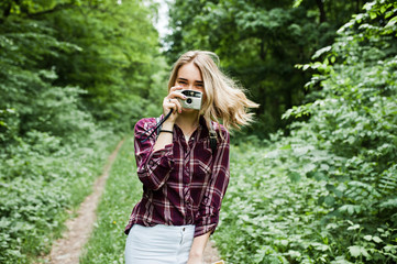 Portrait of a gorgeous young girl in tartan shirt taking pictures with camera in the forest.