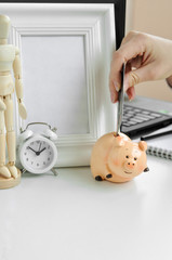 A woman's hand puts credit card in a piggy Bank on the background of a laptop, a clock, a frame, a notebook and a wooden figure of a man at home.
