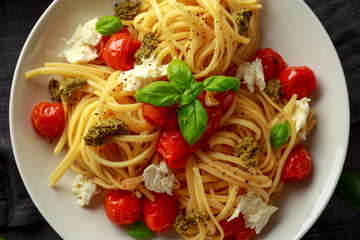 Pasta with green pesto sauce, roasted cherry tomatoes and mozzarella cheese in white plate on dark rustic background