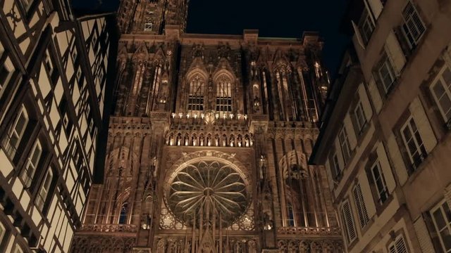 Reverse push-out camera tilt up from base to tower of lighted Notre Dame Cathedral in Petite France old town at night between buildings in Strasbourg, France