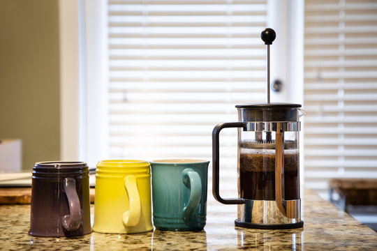 A French Press On A Kitchen Counter Top With Coffee Brewing And Multi Colored Coffee Mugs.