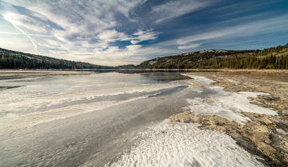 Shoreline of a high mountain lake in the winter with with ice and a green forest