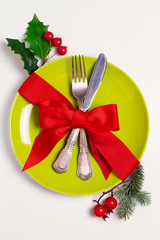 Christmas composition with plate, cutlery, pine branches, ribbon and red berries on white table. Winter holidays and festive background. Christmas eve dinner, New Year food lunch. View from above, top