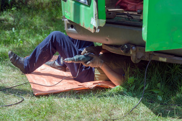 The mechanic lies under the car and repairs the body of a green minibus in the summer on the green grass outside.