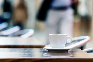 A cup of espresso coffee on a wooden table outdoors with people in the background.