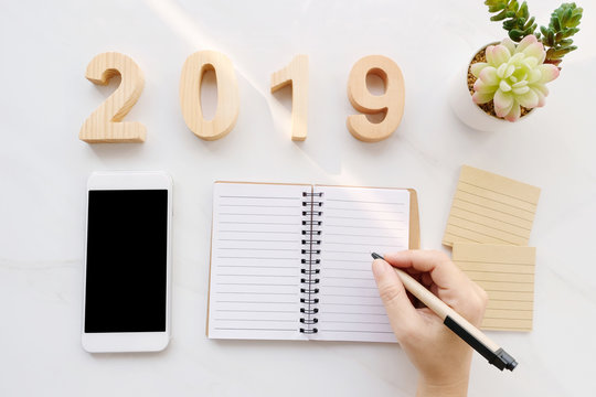 2019 wood letters, hand writing on blank notebook paper, white smart phone with blank screen on white marble table background, 2019 new year mock up, template with copy space for text, top view