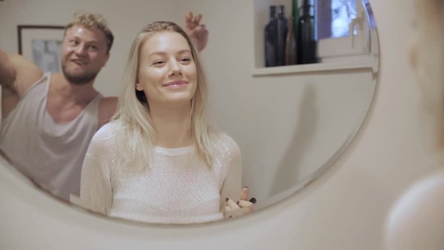 Woman Putting On Her Makeup In The Mirror And Her Boyfriend Jokingly Pops Up Behind To Photobomb Her Making Funny Faces