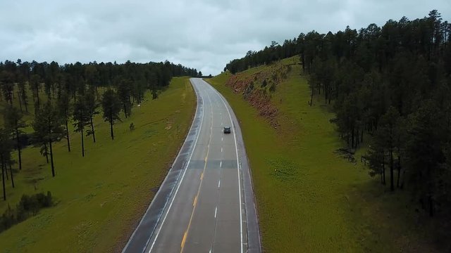 Drone follows silver car moving along concrete highway between wild green forest rocky hills panorama covered with trees