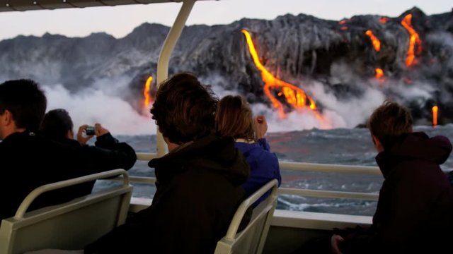 Visitors to Kilauea Hawaii volcanic area watching from boat as red hot lava falls into ocean causing rising steam seen from distance 