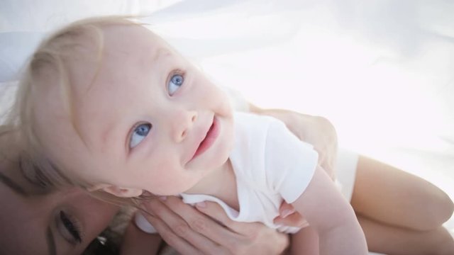 Portrait of cute blonde young Caucasian mother with her baby boy relaxing under the white scarf outdoors 