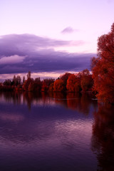 Beautiful landscape with red autumn trees over the lake in perspective and a dramatic sky at sunset