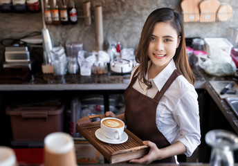 Young beautiful barista wearing brown apron holding hot coffee cup served to customer with smiling face at bar counter